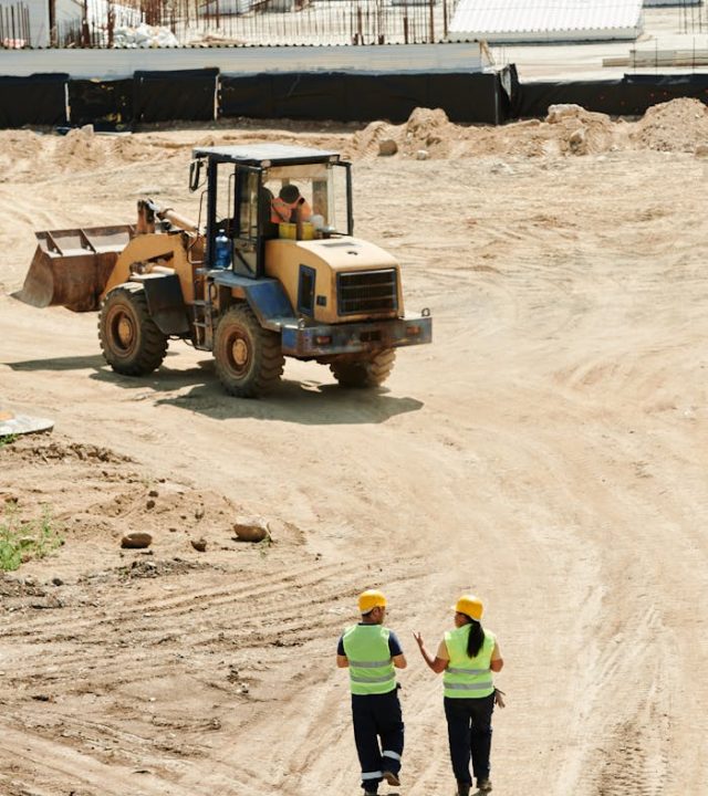 Construction workers in safety gear walking at a busy site with a backhoe in operation.